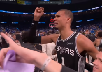A man in a Spurs jersey raises his fist in a stadium full of people.