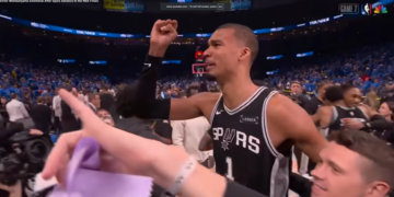 A man in a Spurs jersey raises his fist in a stadium full of people.