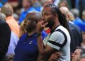 Larry Fitzgerald, a football player, wearing a gray t-shirt with black bands, looking on with his hand on his chin while watching a game with another man.