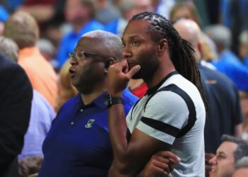 Larry Fitzgerald, a football player, wearing a gray t-shirt with black bands, looking on with his hand on his chin while watching a game with another man.