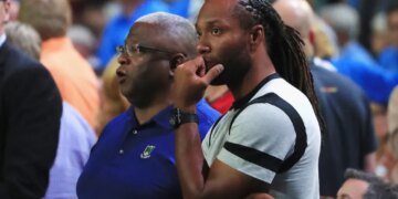 Larry Fitzgerald, a football player, wearing a gray t-shirt with black bands, looking on with his hand on his chin while watching a game with another man.