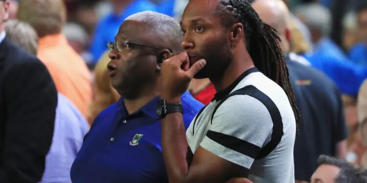 Larry Fitzgerald, a football player, wearing a gray t-shirt with black bands, looking on with his hand on his chin while watching a game with another man.