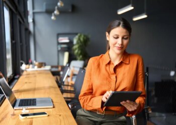 A young Hispanic woman sitting at a desk and using a tablet in a modern office.