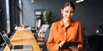 A young Hispanic woman sitting at a desk and using a tablet in a modern office.