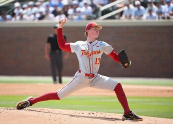 A baseball pitcher wearing a gray uniform with "Trojans" and "13" in red, and red sleeves, pants, and socks, throws a ball on a sunny day.