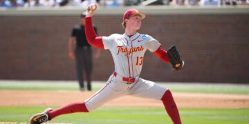 A baseball pitcher wearing a gray uniform with "Trojans" and "13" in red, and red sleeves, pants, and socks, throws a ball on a sunny day.