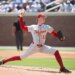 A baseball pitcher wearing a gray uniform with "Trojans" and "13" in red, and red sleeves, pants, and socks, throws a ball on a sunny day.