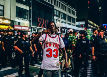 A New York Knicks fan in a jersey making a hand gesture, surrounded by police officers and other fans outside Madison Square Garden at night.