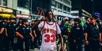 A New York Knicks fan in a jersey making a hand gesture, surrounded by police officers and other fans outside Madison Square Garden at night.