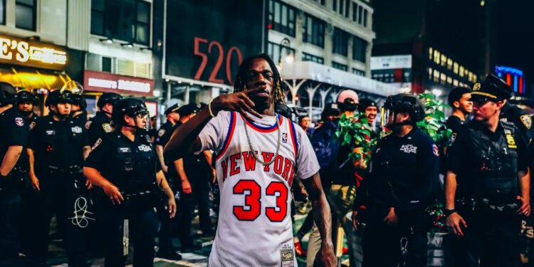 A New York Knicks fan in a jersey making a hand gesture, surrounded by police officers and other fans outside Madison Square Garden at night.