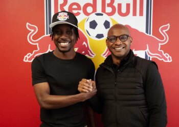 Two men, one in a Red Bull cap, shake hands and smile in front of a Red Bull logo.