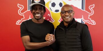 Two men, one in a Red Bull cap, shake hands and smile in front of a Red Bull logo.