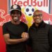 Two men, one in a Red Bull cap, shake hands and smile in front of a Red Bull logo.