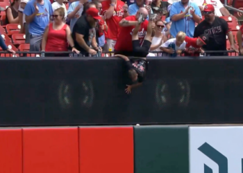 A man holds a child upside down over a stadium wall to retrieve a baseball.