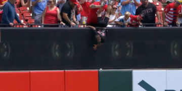 A man holds a child upside down over a stadium wall to retrieve a baseball.