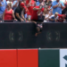A man holds a child upside down over a stadium wall to retrieve a baseball.