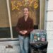 Seattle Mayor Katie Wilson laughs next to a trash can with a business displaying "KATY'S" in the background.