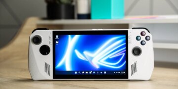 The Asus ROG Ally PC gaming handheld in white standing upright on a wood desk and showing a Windows homescreen.