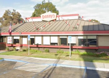 Exterior view of a Denny's restaurant with a red and white sign on the roof, an American flag, and "Open 24/7" banner.