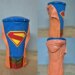 A Supergirl movie drink cup with a blue cup, Superman logo, gold belt buckle, and orange jacket.