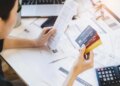 Woman holding multiple credit cards and a long receipt, surrounded by bills and a calculator, appearing concerned about debt.