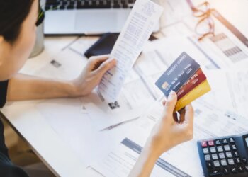 Woman holding multiple credit cards and a long receipt, surrounded by bills and a calculator, appearing concerned about debt.