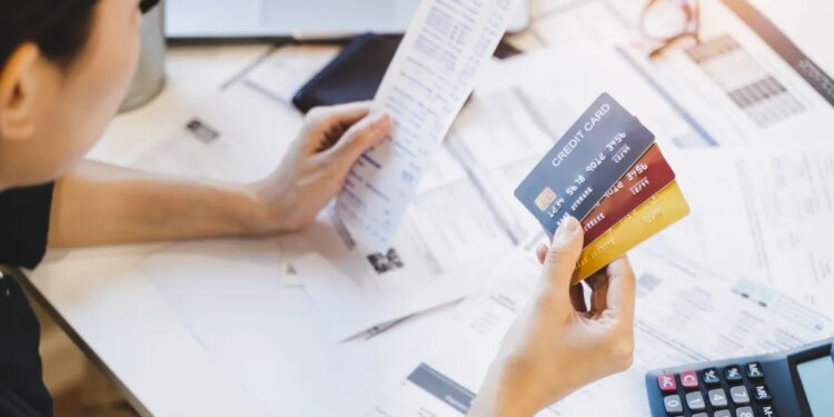 Woman holding multiple credit cards and a long receipt, surrounded by bills and a calculator, appearing concerned about debt.