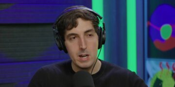 A man wearing headphones and a black shirt speaking into a microphone, with colorful digital art in the background.