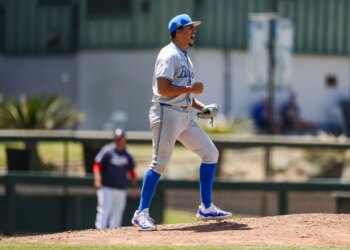 A baseball player on the mound, dressed in a blue cap, grey uniform, and blue socks, cheers with his mouth open and fist clenched.
