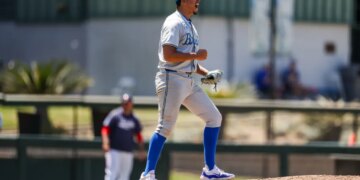 A baseball player on the mound, dressed in a blue cap, grey uniform, and blue socks, cheers with his mouth open and fist clenched.