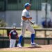 A baseball player on the mound, dressed in a blue cap, grey uniform, and blue socks, cheers with his mouth open and fist clenched.