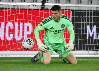 New York City FC goalkeeper Matt Freese in a lime green uniform, kneeling on the grass with a soccer ball to his left.