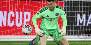 New York City FC goalkeeper Matt Freese in a lime green uniform, kneeling on the grass with a soccer ball to his left.