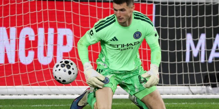 New York City FC goalkeeper Matt Freese in a lime green uniform, kneeling on the grass with a soccer ball to his left.
