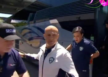 A bald man wearing a white and navy jacket with a UFA logo walking past a security guard and a bus.