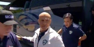 A bald man wearing a white and navy jacket with a UFA logo walking past a security guard and a bus.