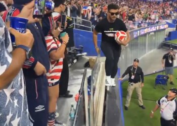 Man in black shirt and sunglasses holding a soccer ball walking across the top of a barrier with security guards nearby.