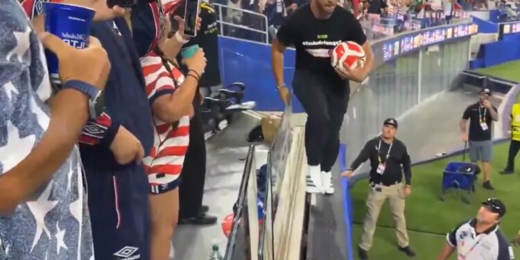 Man in black shirt and sunglasses holding a soccer ball walking across the top of a barrier with security guards nearby.