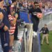 Man in black shirt and sunglasses holding a soccer ball walking across the top of a barrier with security guards nearby.