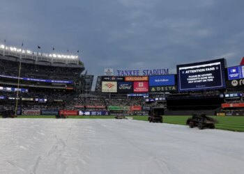 Yankee Stadium field covered by a tarp, indicating a rain delay, with stadium lights on and scoreboards displaying an announcement that the game will not start on time.