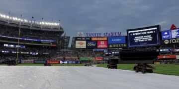 Yankee Stadium field covered by a tarp, indicating a rain delay, with stadium lights on and scoreboards displaying an announcement that the game will not start on time.