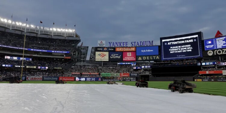 Yankee Stadium field covered by a tarp, indicating a rain delay, with stadium lights on and scoreboards displaying an announcement that the game will not start on time.