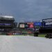 Yankee Stadium field covered by a tarp, indicating a rain delay, with stadium lights on and scoreboards displaying an announcement that the game will not start on time.