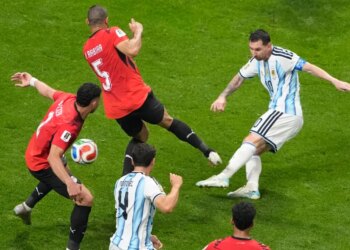 Lionel Messi takes a shot during a soccer match against an opposing team wearing red jerseys.
