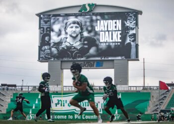 Football players practice on a field under a scoreboard displaying a tribute to late Roughriders player Jayden Dalke.