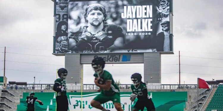 Football players practice on a field under a scoreboard displaying a tribute to late Roughriders player Jayden Dalke.