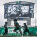 Football players practice on a field under a scoreboard displaying a tribute to late Roughriders player Jayden Dalke.