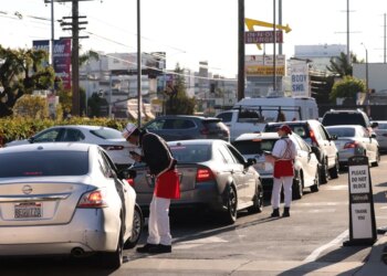 Workers in In-N-Out Burger uniforms take orders from a line of cars at a drive-through.