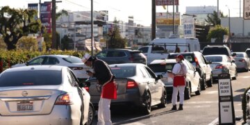 Workers in In-N-Out Burger uniforms take orders from a line of cars at a drive-through.