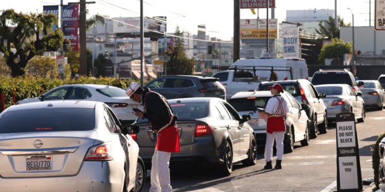 Workers in In-N-Out Burger uniforms take orders from a line of cars at a drive-through.